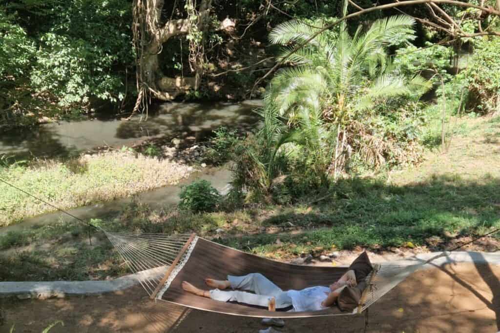 Relaxing woman lying in hammock near river in lush tropical forest, peaceful outdoor scene.