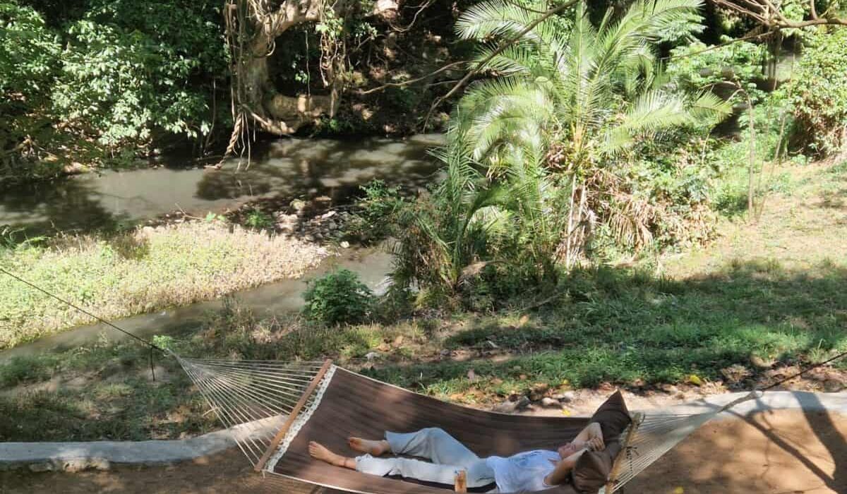 Relaxing woman lying in hammock near river in lush tropical forest, peaceful outdoor scene.